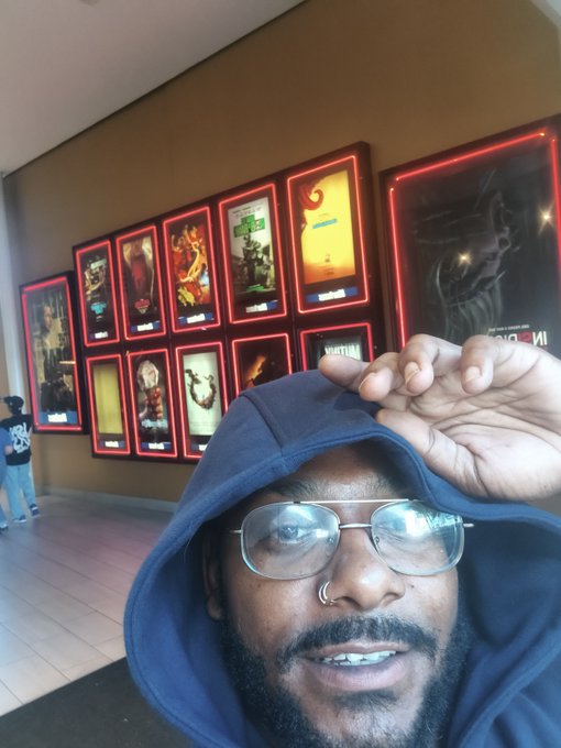 A selfie of a man in a blue hoodie and glasses at a cinema, with multiple glowing red movie poster displays visible on the wall behind him.