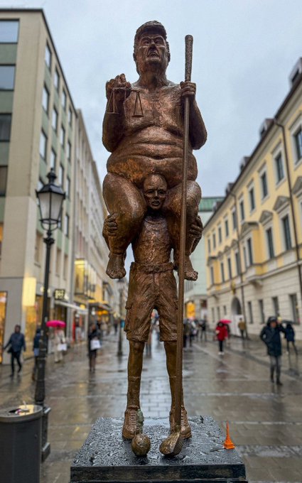 A bronze sculpture depicts a child carrying an overweight man holding a staff and a scale, standing on a pedestal in a European-style plaza with pedestrians and umbrellas in the background. The image visually satirizes financial "plague" themes, portraying a weak man being supported by a child—mirroring the post's critique of a struggling cryptocurrency that needs to be carried by others to reach higher value.