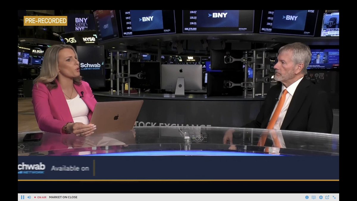 The image shows a man and woman sitting at a table, with a laptop open between them. They appear to be engaged in conversation or discussing something related to the stock market. The background features multiple TVs displaying financial information, indicating that they are likely in a newsroom or a broadcast studio setting.