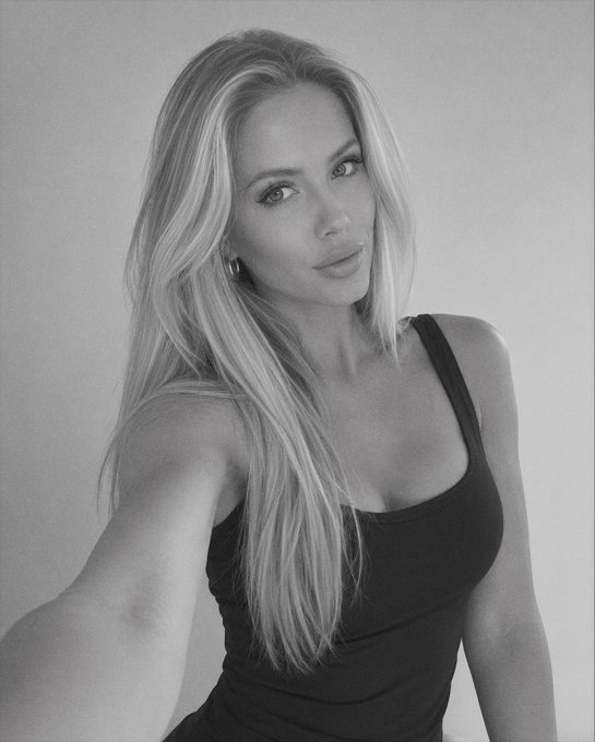 A black and white portrait of a blonde woman wearing a black tank top, taking a selfie against a plain wall with a soft, filtered look.