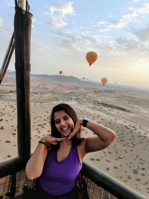 A woman takes a selfie in a hot air balloon basket with multiple colorful balloons visible across the desert landscape at sunrise. The image ties to her post as it showcases her travel experiences, highlighting her outgoing and adventurous spirit as she looks to network at the Token2049 conference.
