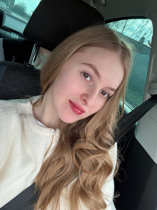 A young woman with long blonde hair and pink blush takes a selfie in a car, wearing a white cardigan with a seatbelt visible across her chest.