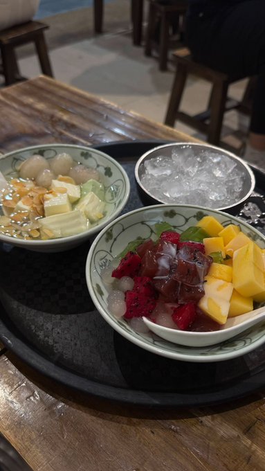 A tray with three bowls of food on a wooden table, including a noodle dish with tofu, a fruit dessert with mango and strawberries, and a small bowl of ice.