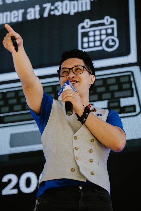 A man wearing glasses, a grey vest, and a blue shirt speaks into a microphone and points upward at an event, with a screen behind him displaying 'at 7:30pm'.