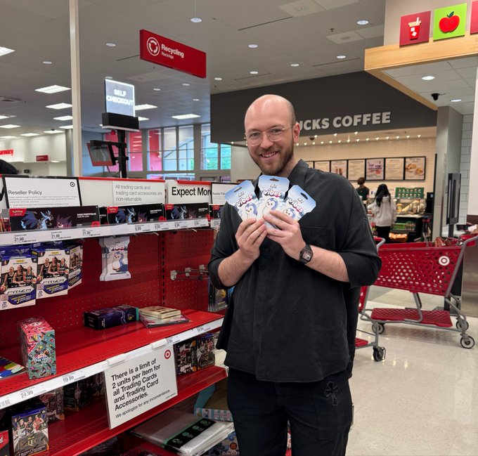 A man wearing glasses stands in front of a store display, holding three small packages or bags. The items are placed on a red shelf that also has other products displayed around him. In the background, there's another person who appears to be shopping as well. A sign is posted near the man with his purchases, which reads "There is an item for sale at Target".