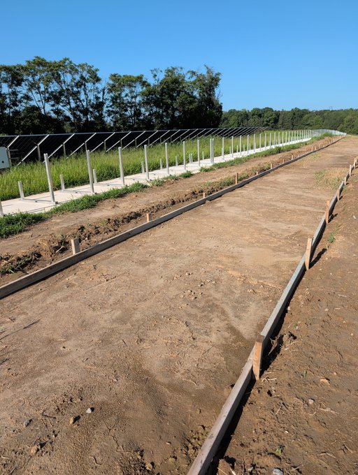 A dirt pathway runs alongside rows of solar panels at what appears to be a solar farm installation. The fenced enclosure with metal posts suggests a developing renewable energy site, fitting the agricultural/industrial theme of the post about a day at the farm.