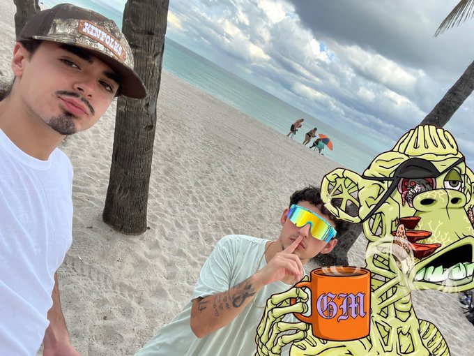 Two young men pose for a selfie on a beach with palm trees and the ocean in the background, overlaid with a Pepe the Frog character holding an orange mug labeled 'GHM.'