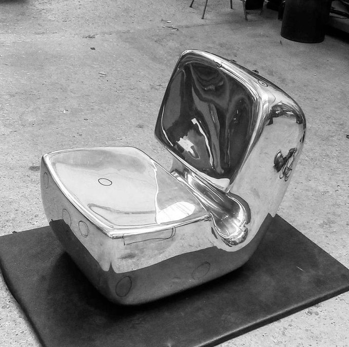 A black-and-white photograph of a polished, reflective silver sculpture shaped like a stylized chair or figure with a curved backrest, displayed on a dark platform.