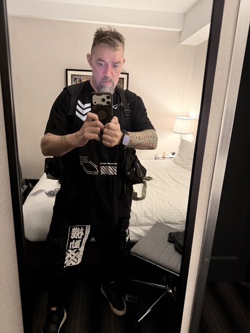 A man with a beard and tattoos takes a mirror selfie in a hotel room, wearing a black t-shirt and backpack while holding a smartphone with a PopSocket.