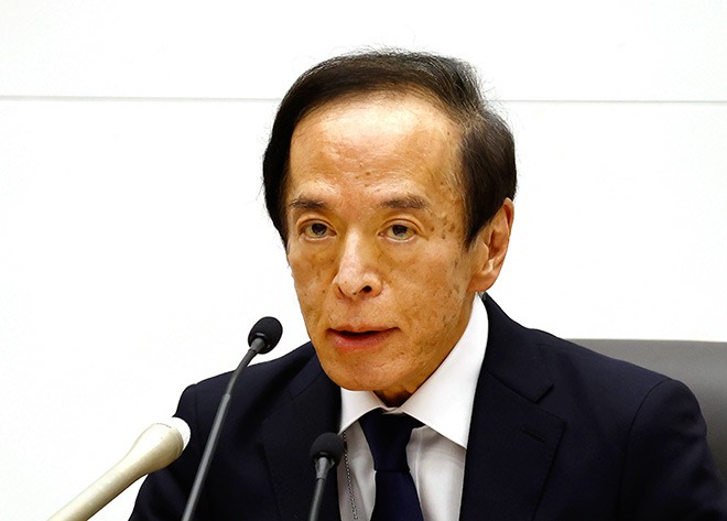The image shows an older man wearing a suit and tie speaking into a microphone at a podium. The man appears to be delivering a speech or addressing an audience, possibly during a press conference or event. The setting seems to be indoors with a white wall in the background.