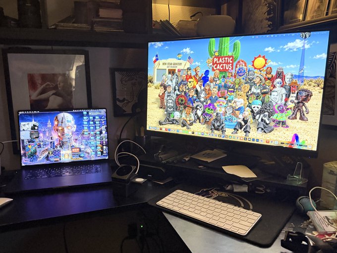 A desk setup featuring a MacBook displaying a colorful skull artwork alongside a larger monitor showing a desert scene with 'PLASTIC CACTUS' and 'NEW YEAR 200 MINT FOR THEM WIZARDS' text, accompanied by an Apple Magic Keyboard.