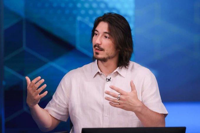 A man with long dark hair and a light pink shirt gestures expressively while speaking during what appears to be a TV interview or financial segment, with a blue geometric-patterned studio background behind him. The image complements the post's discussion of Robinhood ($HOOD) and the impressive early transaction volume on the newly launched Robinhood Chain following their UK Crypto event.
