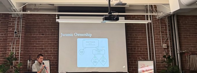 A man speaks at a presentation in a brick-walled room, with a slide titled 'Jurassic Ownership' projected on the screen behind him showing a flowchart diagram.