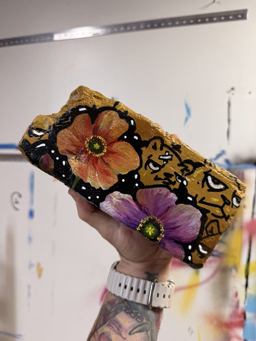 A hand holding up a painted brick featuring floral designs and a stylized face with multiple eyes, with a tattooed wrist and a watch visible.