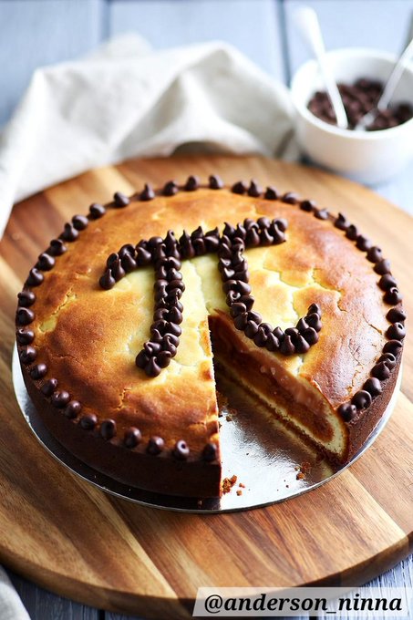 A round cake sits on a wooden cutting board, decorated with chocolate chips piped into a heart pattern on top, with a slice removed. A small bowl of chocolate chips and a linen cloth are in the background, and the image is credited "@anderson_ninna".