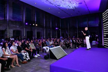 A speaker presents to a large seated audience in a modern, dimly lit venue with purple stage lighting, capturing the energetic atmosphere of one of Circle & Arc's five Istanbul events. The packed room of attendees reflects the diverse gathering of builders, founders, banks, exchanges, and investors mentioned in the post.