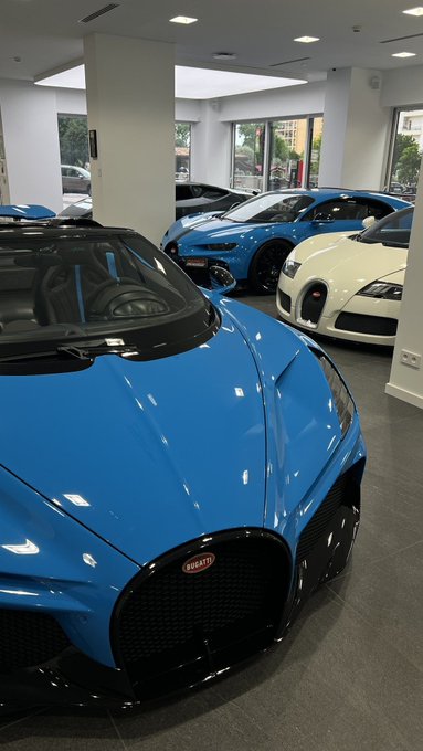 A blue Bugatti sports car is parked prominently in a showroom, with a white Bugatti and another blue car visible in the background.
