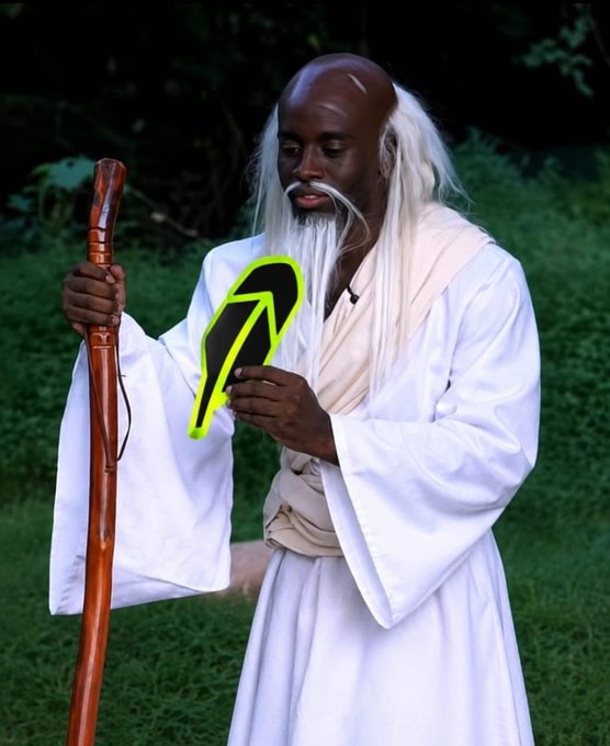 A costumed figure portraying an elderly sage with long white hair, a beard, and white robes, holding a wooden staff in one hand and a black-and-yellow mask in the other, standing on grass.