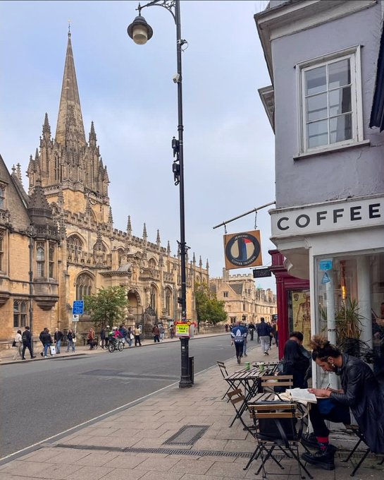 A street scene showing a Gothic-style stone cathedral in the background, with a coffee shop and people sitting at outdoor tables in the foreground.