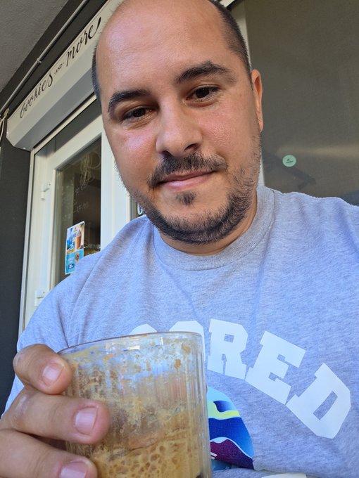 A man with short dark hair and facial hair, wearing a gray t-shirt, smiles while holding a glass of frothy beverage in front of a storefront window.