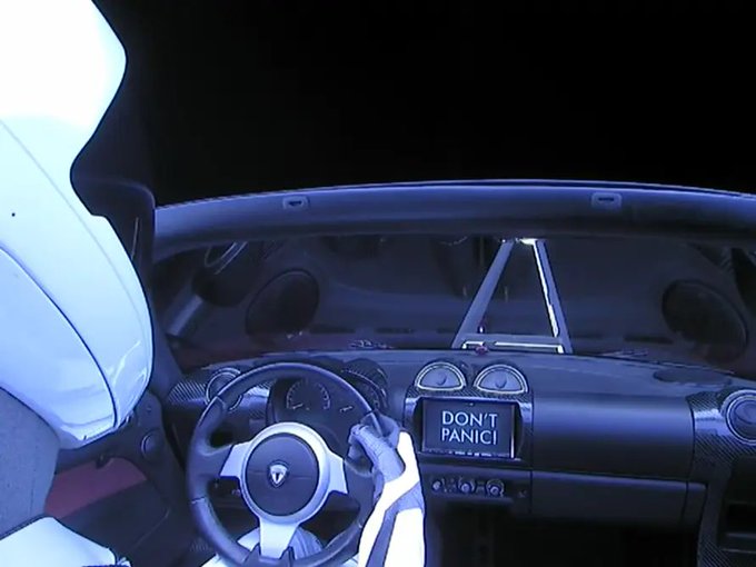 The interior of a car is shown from the driver's perspective, with the dashboard screen displaying the message 'DON'T PANIC!'.