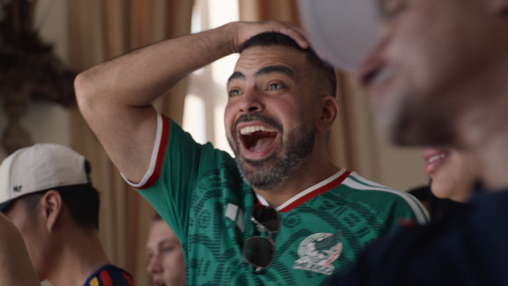 A man in a green Mexico national soccer team jersey is laughing joyfully with his hand on his head in a crowd setting.