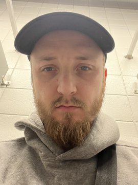 A man wearing a dark cap and gray hoodie taking a selfie, showing a serious expression with a reddish beard.