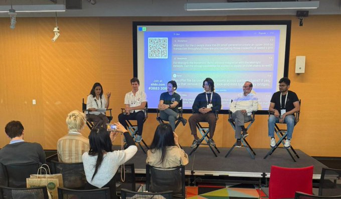 A panel of six speakers seated on stage in front of a presentation screen displaying a QR code, with audience members visible in the foreground.