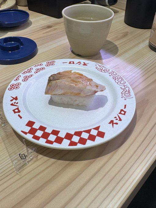 A single piece of nigiri sushi served on a white plate with red Japanese text and checkered patterns (Sushi-ro logo), accompanied by a cup of tea and a blue lid on a light wood counter, likely at a conveyor belt sushi restaurant.