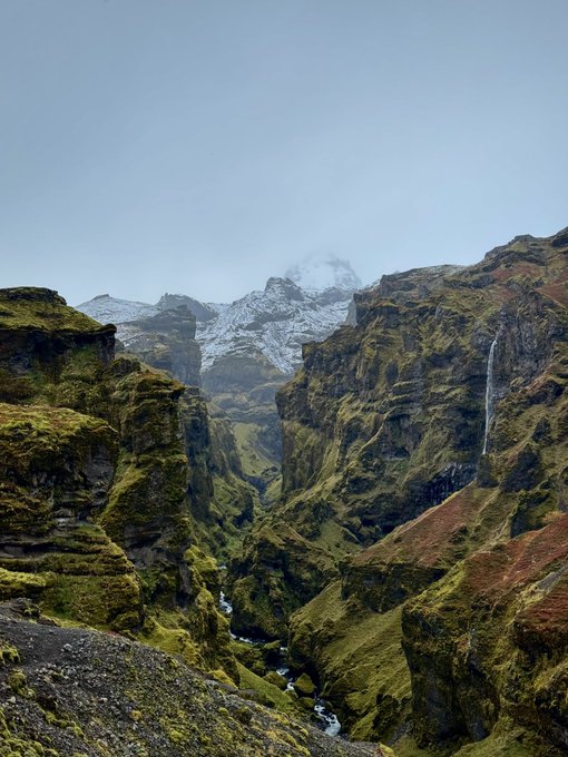 A scenic canyon with moss-covered rocky walls frames a narrow gorge, with snow-capped mountains and a waterfall visible in the distance under a clear sky.