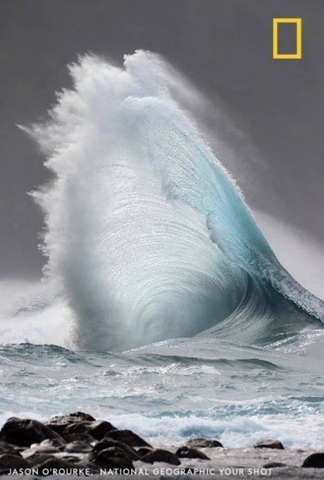 A dramatic photograph of a large turquoise ocean wave cresting and crashing, credited to Jason O'Rourke, National Geographic Your Shot, with the National Geographic logo in the corner.