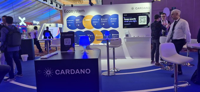 A trade show booth for Cardano features the company name and ecosystem partners, with attendees networking in the background.