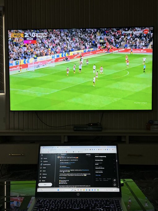 A large TV screen shows a live Premier League match with HUL 2-0 MUN at the 48:22 mark, while an HP laptop below displays a Twitter thread discussing Manchester United's poor start to the season, tying together the visual of the lopsided score with the post's lament about United's inability to compete for the Premier League title.