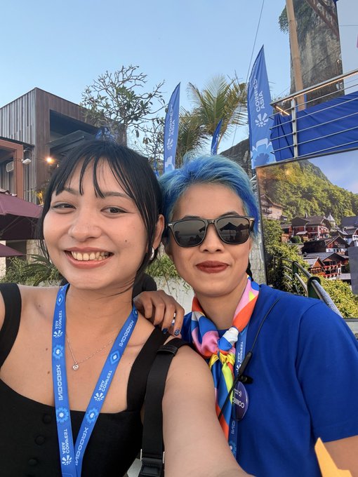A selfie of two women at an outdoor event with BNB Chain lanyards, with one sporting bright blue hair and sunglasses, posing against a backdrop of flags and buildings.