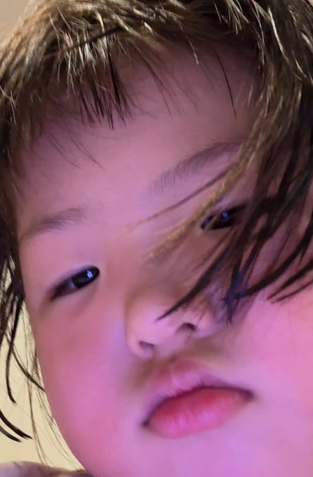 A close-up photograph of a young child's face with hair partially covering their eyes, illuminated with a pinkish hue.