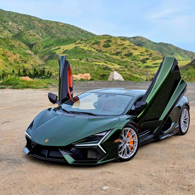 A dark green Lamborghini Revuelto with its scissor doors open is parked on a dirt road with grassy hills in the background.