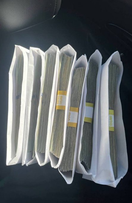 Several bundles of cash wrapped in bank deposit bands are stacked on a car seat, with visible labels reading "$10,000" — implying the person is making money, likely referring to weekly earnings in cash.