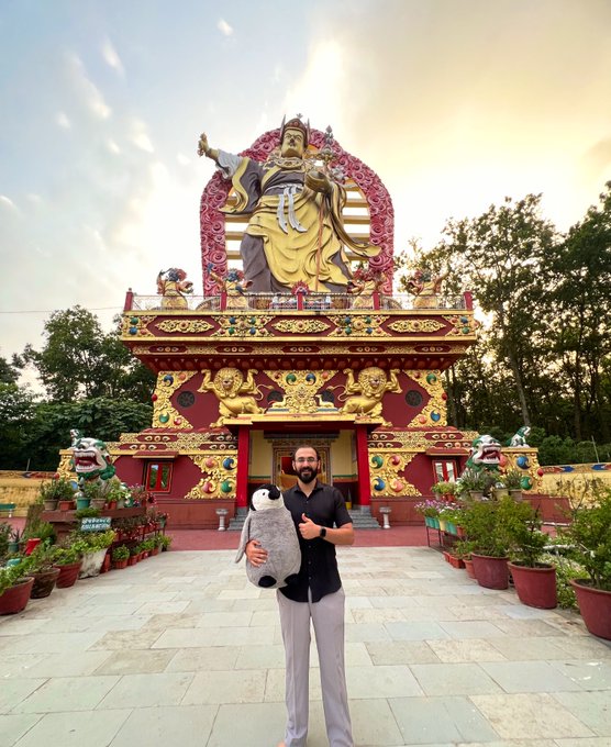 A man holding a large stuffed owl stands in front of an ornate, colorful Buddhist temple featuring a large golden statue of a deity.