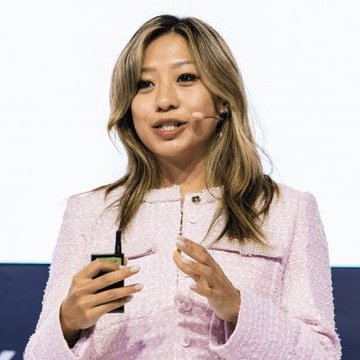 A woman with shoulder-length brown hair wearing a light pink tweed jacket, holding a small black device and appearing to speak at an event.
