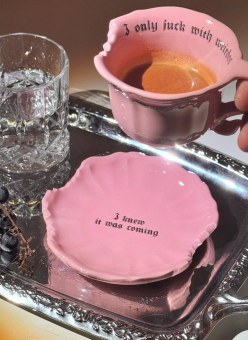 A pink ceramic teacup and saucer set with gothic text reading 'I only fuck with wealthy' and 'I knew it was coming,' filled with coffee and displayed on a silver tray.