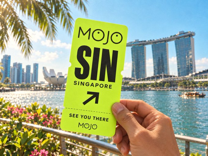A hand holds a bright yellow 'MOJO SIN SINGAPORE' branded ticket/pass in front of Singapore's Marina Bay skyline, featuring the iconic Marina Bay Sands and ArtScience Museum.