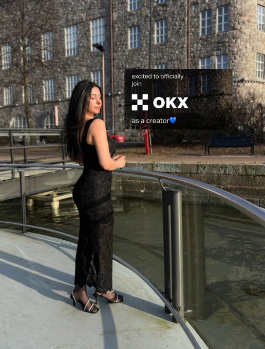 The image features a woman standing next to a metal railing with her back to the camera. She is wearing a black dress and high heels, giving off a stylish and elegant vibe. The background of the photo shows an outdoor setting near water, possibly a river or canal. There are also some buildings visible in the scene.

The image includes text that reads "angels i have a big news i’m officially an @okx creator started by filming videos in my room, explaining crypto with fries some laughed at the analogies, some told me to be more serious, less girly i kept the fries and the slay yesterday i signed as an official". This text is related to the woman's post on social media.