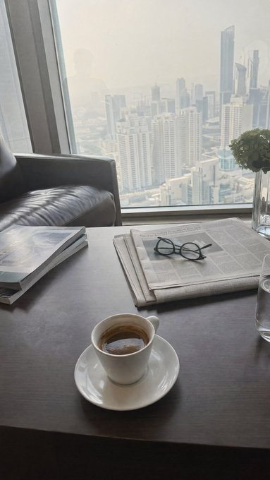 A small espresso cup sits on a saucer on a wooden table, with reading glasses resting on a folded newspaper, a black leather chair, and a window overlooking a hazy city skyline in the background.