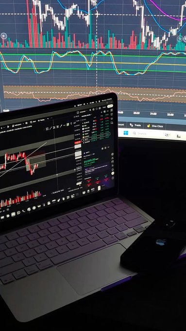 A laptop displaying trading charts and data sits on a desk, with a larger monitor in the background also showing financial charts and graphs.