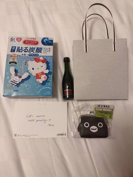 A photo of merchandise items laid out on a white sheet, including a Hello Kitty face mask package, a small bottle, a gray paper bag, a handwritten card, and a black character pouch in plastic packaging.