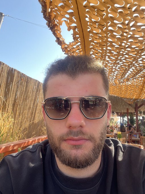 A man with a beard and dark sunglasses takes a selfie outdoors, sitting beneath a decorative laser-cut wooden canopy that casts intricate shadow patterns, with bamboo fencing and other patrons visible in the background.