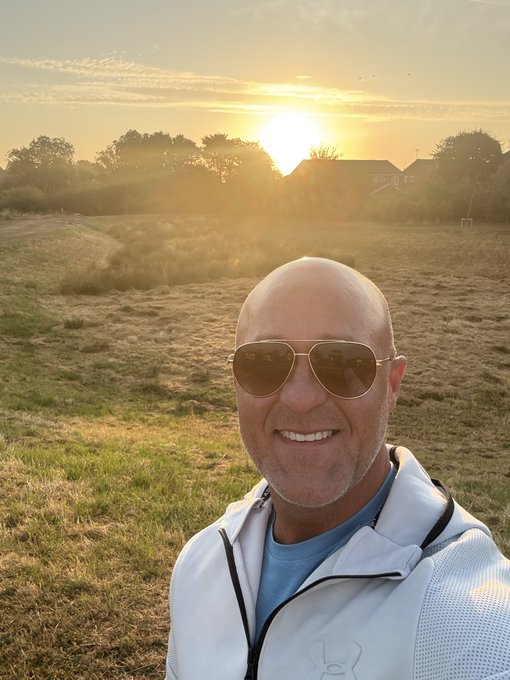 A selfie of a bald man wearing aviator sunglasses and a light-colored jacket with a bunny logo, standing in a grassy field at sunset.