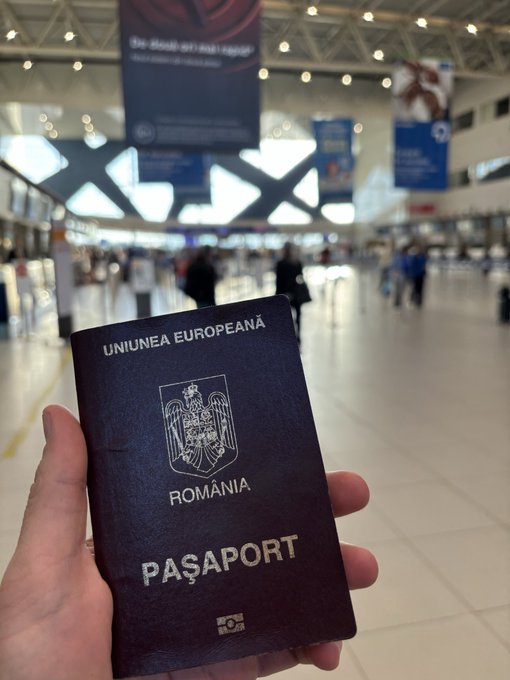 A hand holds a Romanian passport (UNIUNEA EUROPEANĂ ROMÂNIA PAȘAPORT) in an airport terminal with banners overhead.