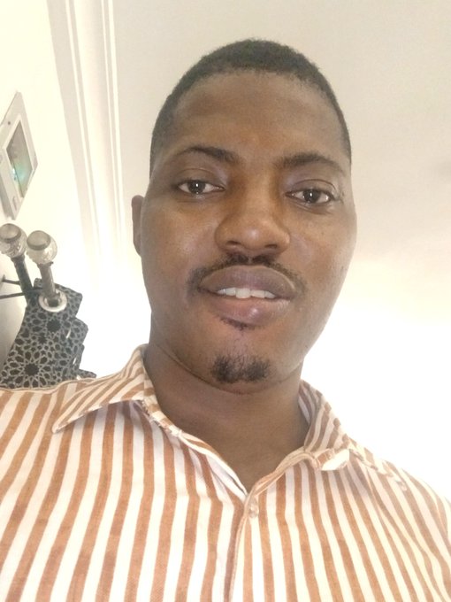 A close-up selfie of a young man wearing a brown and white vertically striped collared shirt, indoors with some electronics visible in the background.