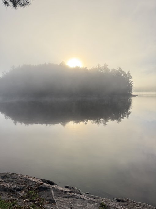 A serene landscape photograph of a misty lake at sunrise or sunset, with a forested island silhouetted against the glowing sun and its reflection mirrored on the calm water, framed by rocky shore in the foreground.